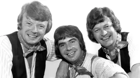 A black and white publicity image of the band, all three are wearing red neckerchiefs and white shirts and smiling