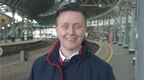 Jess Mawinney-Coates is standing on a platform at Newcastle station. She's a young woman with a big smile, short hair and blue eyes. 