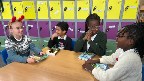 Four children sit around a wooden classroom table holding books. Behind them is a wall of colourful lockers in yellow, purple, and green. One child is wearing a festive headband with red antlers, and the books on the table include various reading materials.