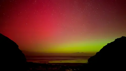 Shedders/Weather Watchers A red and yellow sky is studded with stars above a beach and reflected in the sea and wet sand with silhouetted cliffs left and right framing the view.