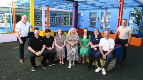 A group of people sitting or standing in an area that is surrounded by blue walls and colourful columns. They include men from left to right Mark Wellings from UK Electrical, Tim Lewis, David Cotterill, both Inco Contracts and Myles Weldon DMW.  Females - Louise Skyte, Jo Catchpole, both Inco Contracts, Amanda Winwood, Amie Rogers and Kirsty Lewis,all Your RWTC.


