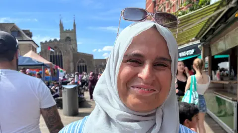 Jenny Kirk/BBC A head and shoulders image of a woman wearing a headscarf with glasses on her head. She is standing in a city centre square and looking into the camera smiling. Behind him are stalls and people walking around. 