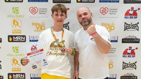 RYAN ASHWORTH A boxer (left of pic) stands with his coach. The boxer has a gold medal around his neck, is wearing a white T-shirt and is holding a certificate. The coach has a beard, is wearing a white T-shirt and is holding his left fist to the camera.