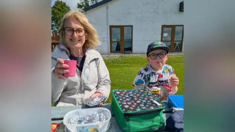 Jane Richards Helen sat with her grandson on a picnic bench in the sun. They are both eating a packed lunch on the table in front of them and smiling. Helen holds a pink plastic cup in the air, she is wearing a grey raincoat. 