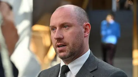 Getty Images a man with close-cropped hair and beard is wearing a grey suit, white shirt and black tie