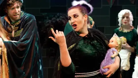 Michelle Parker playing Gothel holding a baby and looking over her shoulder