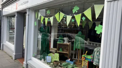 The front of the Save the Children charity shop in Ballycastle. It has green bunting and shamrocks on the window. Green items of clothing including a dress, a St Patrick's Day hat, children's clothes and toys are on display.