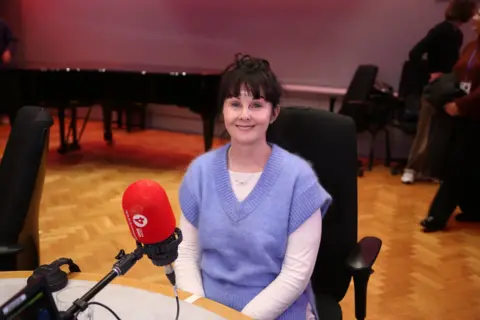 A woman smiles while sitting behind a micrpphone. She wears a light purple vest and has a brown fringe.