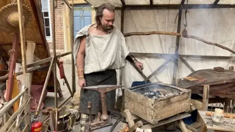 A man dressed as a Roman blacksmith, with a white tunic and a brown apron, tends to smouldering coals in a wooden crate while surrounded with iron-working equipment under an open canvas tent.
