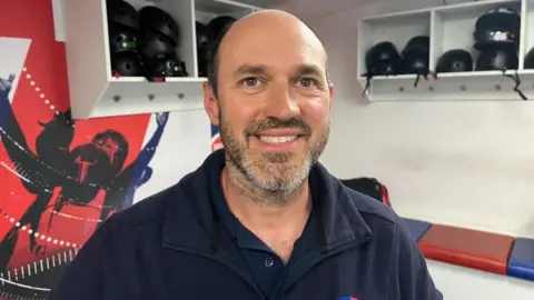 Phil Harris from Young Bristol, photographed in a bobsleigh locker room with helmets on alcoves behind him