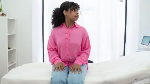 A young woman wearing jeans and a pink shirt sits on a bed waiting for her cervical screening
