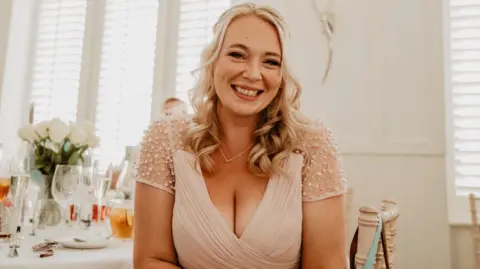 A young woman with long blonde hair wearing a white dress smiling at the camera with a table laid out with glasses and flowers in the background.