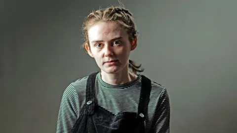 Stephanie stands against a gray backdrop and looks directly on the camera. Her hair is in plaque and wearing a blue dungaras and a stripy jumper.