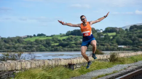 Sam has brown hair and is wearing sunglasses. He is in an orange, blue and white running vest that has the motor neurone disease foundation logo on it and the number 125. He is jumping in the air with both arms outstretched. 