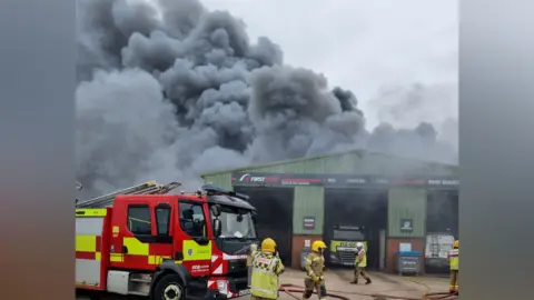 A thick, grey plume of smoke rises from a green, metal industrial warehouse. There is a fire engine parked out front and firefighters walking in front of the building.