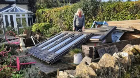 Adrian An image taken in a garden in Condicote. A lady is stood amongst fencing, chairs, and other objects which have been transported by the weather.