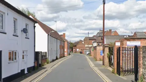 Google A street map view of a narrow street with terraced homes on the left and gates and red brick walls at the entryways to business and industrial buildings on the right.