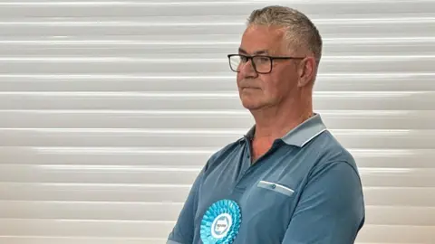A man stands side on to the camera. He wears a blue T-shirt, with a blue Reform UK rosette. In the background is a white shutter and a beige bar. 