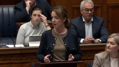 NI Assembly Liz Kimmins speaking in front of a podium in the assembly. She has light brown hair tied back, wearing a navy jacket with gold buttons and khaki top underneath. Other MLAs are in the background.