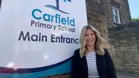 A woman with blonde shoulder-length hair is wearing a striped top and a dark blue jacket. She is stood outside a school next to a sign which says Carfield Primary School Main Entrance.
