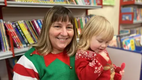 Laura Coffey/BBC Smiling woman with long fair hair wearing a green, white and red Christmas jumper holds a young girl wearing a red sweater
