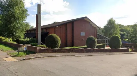 A modern church, made from red brick, with a large structure to the left. There is a small wall round the building, with three round bushes. There are also trees around the building. A road runs down the side of the church. 