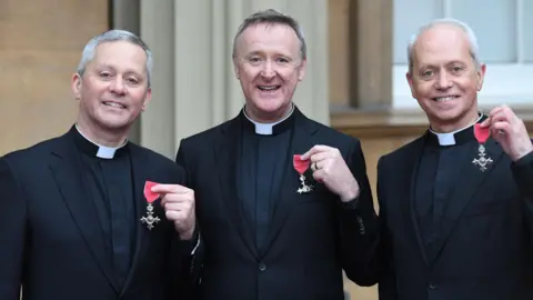 Getty Images Three priests - Fr Martin O'Hagan, Fr David Delargy and Fr Eugene O'Hagan hold up their MBE awards while smiling for the camera outside of Buckingham Palace. They wearing black priest vestments and collars.