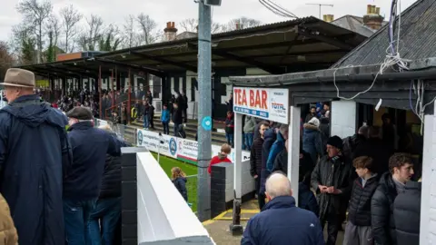 A picture of fans watching Maidenhead United at York Road, with a sign for the tea bar prominent.