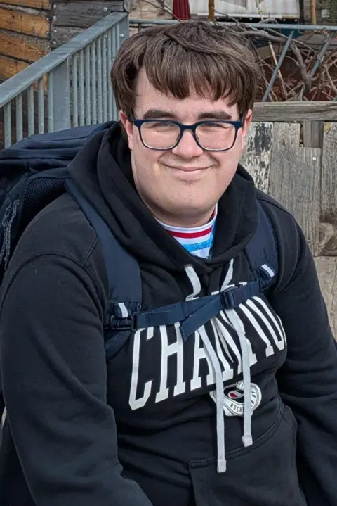 Alexander McLean A man looking at the camera. He wears glasses and has a blue hoody on as well as a blue backpack.