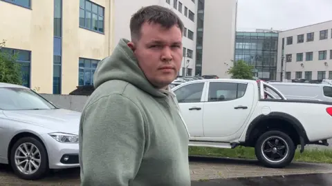 A man wearing a light green hoodie looks at the camera. There are multiple cars parked behind him as well as a cream and white coloured building.
