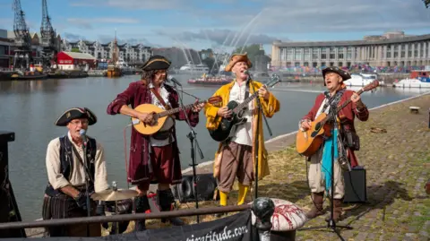 Bristol Harbour Festival Four male musicians, dressed as pirates, stand in front of mics singing and playing guitars, in front of Bristol Harbour Side. There is a large boat in the background spraying jets of water into the air.
