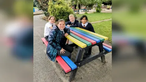 Nunthorpe Primary School Four children smile at the camera while sitting around a rainbow coloured picnic bench