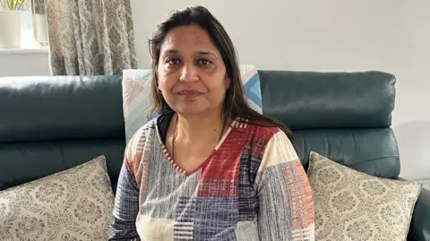 A woman with dark hair and a multiple colour jumper siting on a green sofa inside her living room.