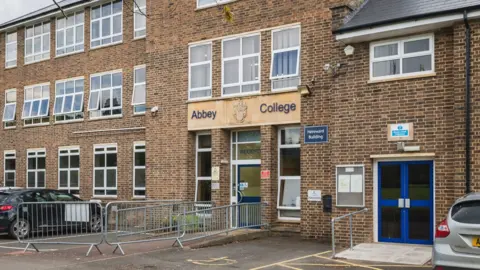 Outside Abbey College. It is a brick building with cars parked outside. Above the blue front entrance door is the school's name and crest.