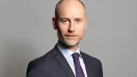 Parlamento del Reino Unido Stephen Kinnock: un hombre barbudo, con barba de color marrón y gris ajustado con camisa pálida, corbata azul y roja y chaqueta gris  