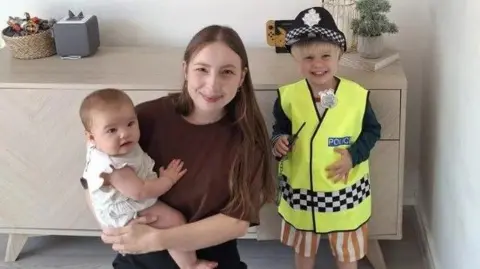 South Yorkshire Police Baby (left), woman with long straight brown hair in a brown top and black trousers (centre), and a blonde three year old boy (right) smiling widely in a police jacket and orange and white striped shorts in front of a living room cabinet