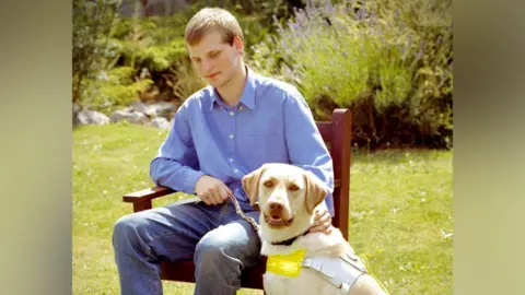 Sean Dilley Sean Dilley as a young man, wearing a blue collared shirt and jeans, sitting in a chair in a garden next to his guide dog Brandy