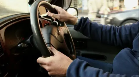 Generic picture of a man behind the wheel of a car. He is wearing a blue jumper. 