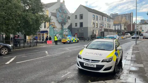 Photo taken near Plymouth's Barbican showing two police cars closing the area. The vehicles are parked on the road with a red "road closed" sign.