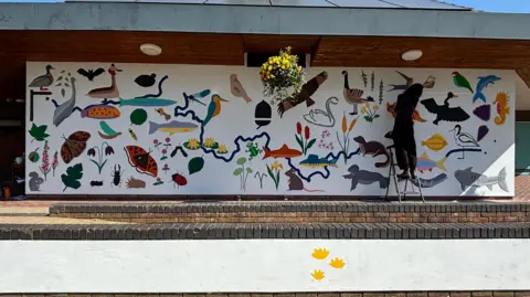 Vickie Amiralis on the right of the image stood on a ladder painting the mural which has a variety of wildlife painted on a white wall.