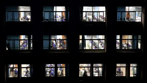 Getty Images A wide shot of a school building at night, with windows to classrooms showing students studying