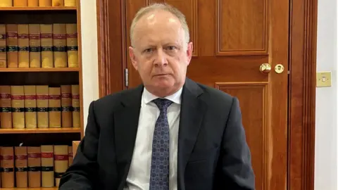 Northern Ireland Office Lord Turnbull, wearing a dark suit and tie, sits in front of a wooden door and a bookcase filled with legal texts