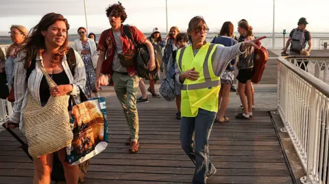 A volunteer gives indication to holidaymakers from Lege-Cap-Ferret upon their arrival in Arcachon by boat during an evacuation operation of the entire Cap-Ferret peninsula. The tourists are seen panicked on a wooden getty while the holidaymaker is in a hi-vis jacket. The sea is seen in the background.