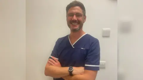 North Bristol NHS Trust Euan Booth in his blue nurses scrubs crossing his arms and smiling
