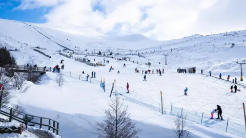Angus Trinder/Cairngorm Mountain Snowsports enthusiasts at Cairngorm Mountain. The skiers and snowboarders are dotted across snow-covered slopes. It is a bright, sunny day.