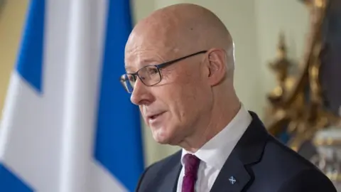 PA Media A bald man with glasses at a press conference. He is wearing a dark suit, white shirt and purple tie, standing side on to the camera in front of a saltire. 