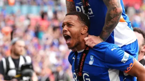 Shaun Hobson wearing an Oldham Athletic jersey and a medal round his neck appears to be shouting, with the arms of James Norwood leaning on his shoulders. In the background are the crowds in Wembley Stadium, but out of focus.