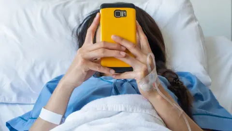 A stock image of a young woman in a hospital bed wearing a blue gown with a yellow mobile phone