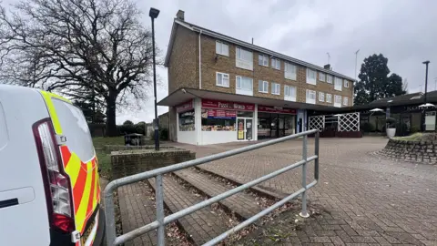 A general view of Priestwood Square in Bracknell, a residential area with two floors of flats above shops, including a takeaway called Pizza World. A police van is parked next to a paved area, with just its boot visible