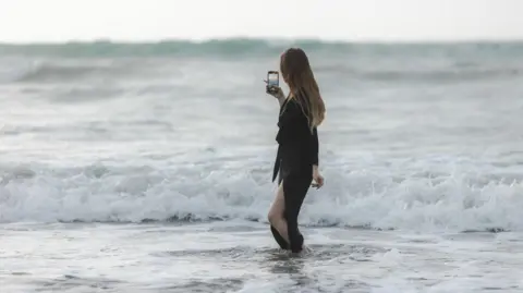 A woman with long dark blonde hair stands in the ocean. She is facing away from the camera and holding her phone in her right hand, she is recording the view of the ocean waves. The woman wears a long sleeved black dress, which has a long side split in the skirt showing her left leg.